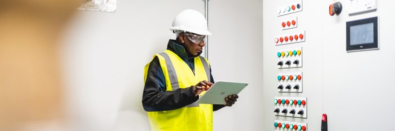 A man in a high vis yellow jacket with a iPad looking at a control panel of buttons A man in a high vis yellow jacket with a iPad looking at a control panel of buttons