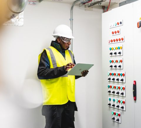 A man in a high vis yellow jacket with a iPad looking at a control panel of buttons A man in a high vis yellow jacket with a iPad looking at a control panel of buttons