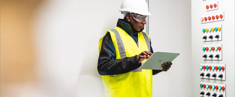 A man in a high vis yellow jacket with a iPad looking at a control panel of buttons A man in a high vis yellow jacket with a iPad looking at a control panel of buttons